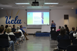 The image shows Donalda signing to the audience with a slideshow image in the background that reads “Why not host the Deaflympics alongside the Olympics and Paralympics?"